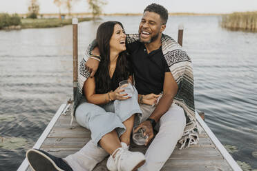 Happy couple drinking wine while sitting on pier against lake - SMSF00435