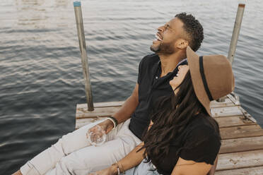 Happy couple drinking wine while sitting on pier - SMSF00447