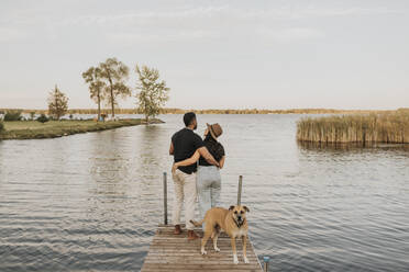 Dog standing by couple on pier against lake - SMSF00449