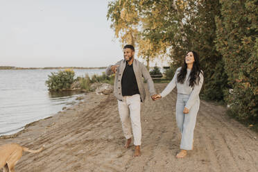 Smiling boyfriend and girlfriend holding hands while walking with dog by lake - SMSF00459