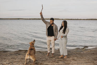 Paar hält Hände beim Spielen mit Hund gegen See während des Sonnenuntergangs - SMSF00460