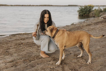 Frau spielt mit Hund, während sie bei Sonnenuntergang am See hockt - SMSF00462