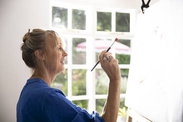 Aktive ältere Frau, die im Stehen zu Hause ein Bild auf Leinwand malt - PMF01483