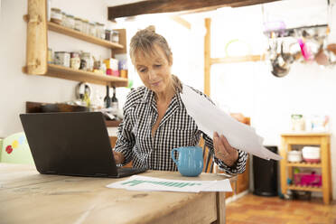 Ältere Frau arbeitet an einem Laptop und hält Papier in der Hand, während sie in der Küche zu Hause sitzt - PMF01487