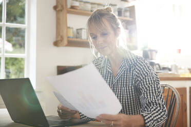 Ältere Frau mit Papier in der Hand sitzt mit Laptop in der Küche zu Hause - PMF01489