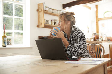 Lächelnde ältere Frau, die wegschaut, während sie Kaffee trinkt und mit einem Laptop in der Küche zu Hause sitzt - PMF01491