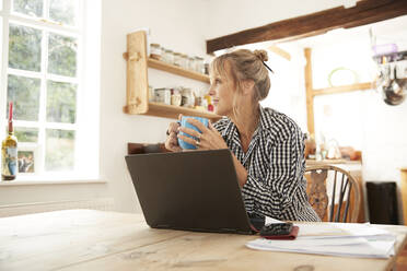 Ältere Frau, die wegschaut, während sie Kaffee trinkt und mit einem Laptop in der Küche zu Hause sitzt - PMF01492