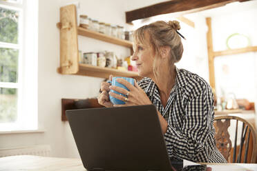 Aktive ältere Frau, die Kaffee trinkt und wegschaut, sitzt mit Laptop in der Küche - PMF01493