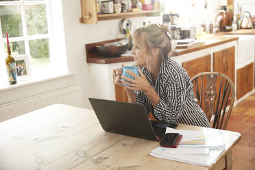 Seniorin hält Kaffeetasse und schaut weg, während sie mit Laptop auf dem Tisch in der Küche zu Hause sitzt - PMF01494
