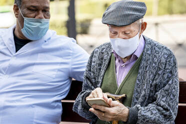 Senior man using smart phone wearing protective face mask sitting with mature male on bench during COVID-19 - GGGF00020