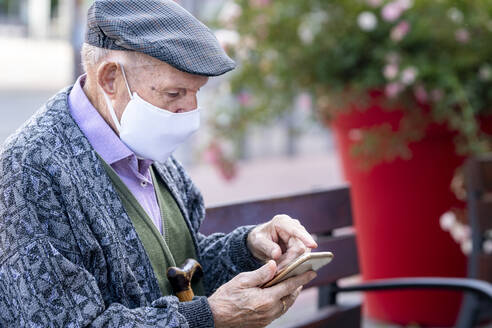 Active senior man using smart phone sitting on bench outdoors - GGGF00021
