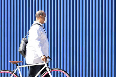Mature man walking with bicycle by blue metal wall on sunny day - GGGF00030
