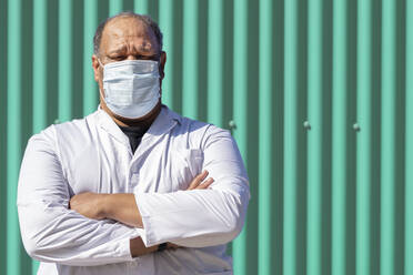 Mature man with arms crossed standing against green metal wall on sunny day - GGGF00031