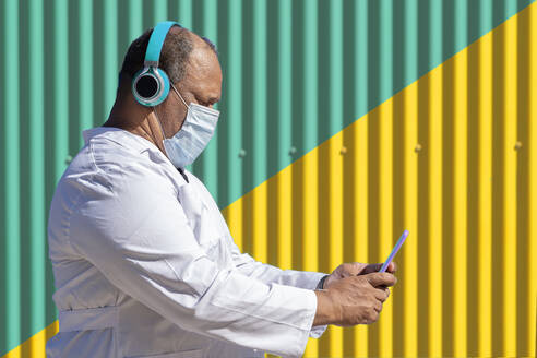 Mature man listening music through headphone while using smart phone standing by green and yellow wall during COVID-19 - GGGF00032