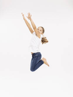 Fröhliches Mädchen springt mit erhobenen Armen vor weißem Hintergrund im Studio - DHEF00507
