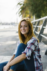 Blond woman sitting on footpath against railing - XLGF00671