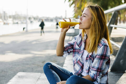 Young woman drinking juice while sitting on footpath in city stock photo