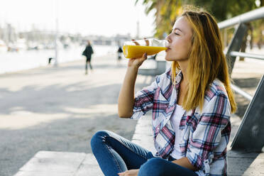 Young woman drinking juice while sitting on footpath in city - XLGF00672