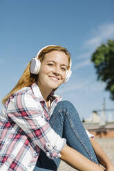 Lächelnde Frau, die mit Kopfhörern Musik hört, sitzt auf einem Fußweg an einem sonnigen Tag - XLGF00677