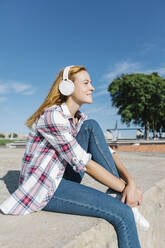 Woman listening music through headphone looking away while sitting on footpath during sunny day - XLGF00679