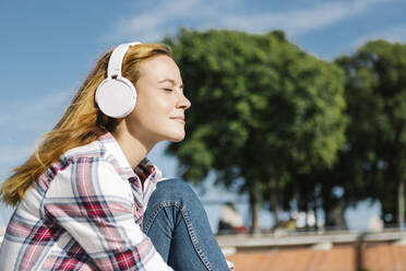 Frau mit geschlossenen Augen hört Musik über Kopfhörer an einem sonnigen Tag - XLGF00681