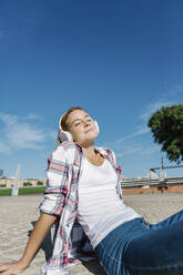 Woman with eyes closed listening music through headphone sitting footpath during sunny day - XLGF00682