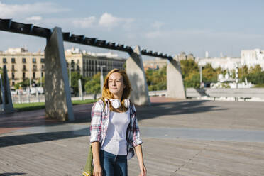Junge Frau mit Kopfhörern um den Hals, die an einem sonnigen Tag auf einem Fußweg spazieren geht - XLGF00684