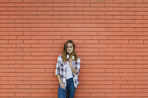 Young woman with headphone around her neck standing against brick wall stock photo