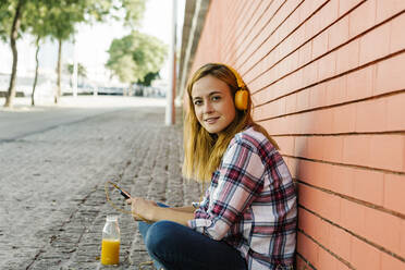 Frau hört Musik auf dem Smartphone über Kopfhörer, während sie an einer Mauer sitzt - XLGF00695
