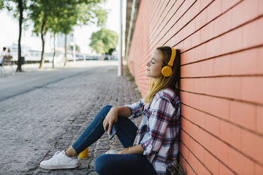 Woman listening music with eyes closed while sitting on footpath against brick wall - XLGF00696