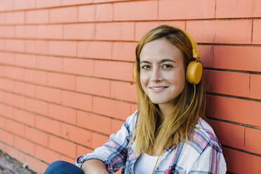 Lächelnde Frau hört Musik über Kopfhörer und sitzt an einer Mauer - XLGF00697