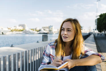 Nachdenkliche junge Frau mit Buch auf dem Fußweg am Meer sitzend - XLGF00702