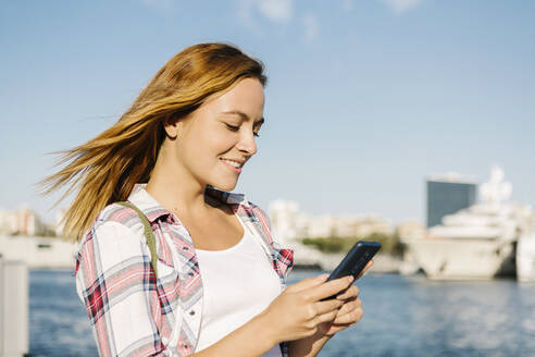 Frau Textnachrichten auf dem Smartphone, während sie an einem sonnigen Tag am Meer steht - XLGF00704