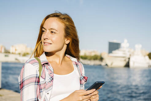 Frau benutzt Smartphone und schaut weg, während sie an einem sonnigen Tag am Meer steht - XLGF00705