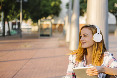 Woman wearing headphone looking away using digital tablet sitting on bench during sunny day stock photo