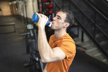 Athlete man drinking water while standing in exercise room - SNF00701