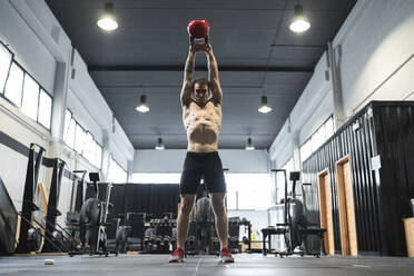 Männlicher Sportler, der im Fitnessraum stehend eine Kettlebell hebt - SNF00729