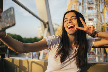 Cheerful woman on Ferris wheel using mobile phone while taking selfie at amusement park - OYF00233
