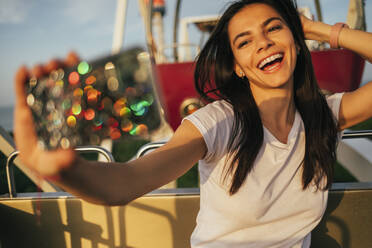 Schöne junge Frau lacht, während sie ein Selfie mit der Hand im Haar auf dem Riesenrad im Vergnügungspark macht - OYF00240