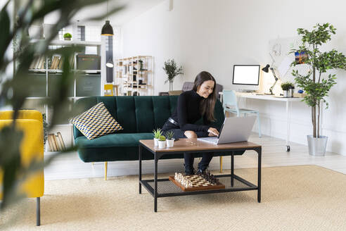 Smiling young female blogger using laptop while sitting in living room at loft apartment - GIOF09409