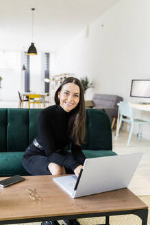 Smiling young female blogger sitting with laptop on sofa at home - GIOF09412