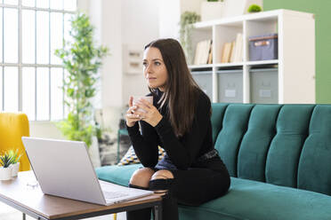 Thoughtful young woman holding coffee cup while sitting with laptop on sofa in loft apartment - GIOF09413