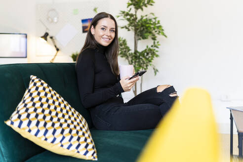 Smiling female blogger holding smart phone and coffee cup while sitting on sofa at loft apartment - GIOF09417