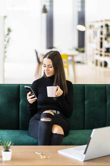 Young female influencer holding smart phone and coffee cup while sitting on sofa at loft apartment - GIOF09418