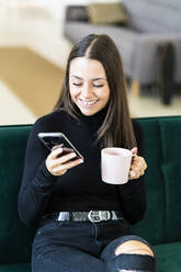 Lächelnde junge Influencerin hält Smartphone und Kaffeetasse, während sie auf dem Sofa in einer Loftwohnung sitzt - GIOF09419