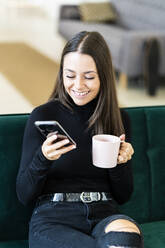 Smiling young female influencer holding smart phone and coffee cup while sitting on sofa at loft apartment - GIOF09419