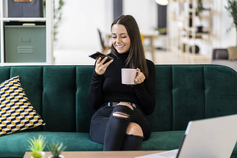 Smiling young female blogger holding smart phone and coffee cup while sitting on sofa at loft apartment stock photo