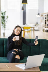 Lächelnde schöne weibliche Influencerin, die ein Selfie mit einer Kaffeetasse macht, während sie zu Hause auf dem Sofa sitzt - GIOF09422