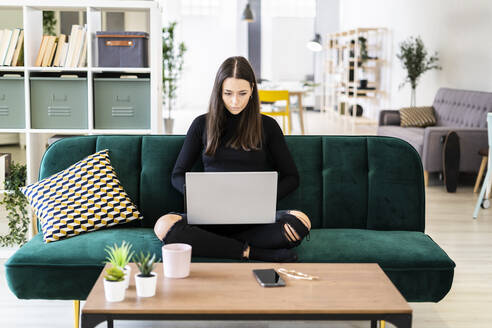 Beautiful young woman using laptop for blogging while sitting on sofa at loft apartment - GIOF09423