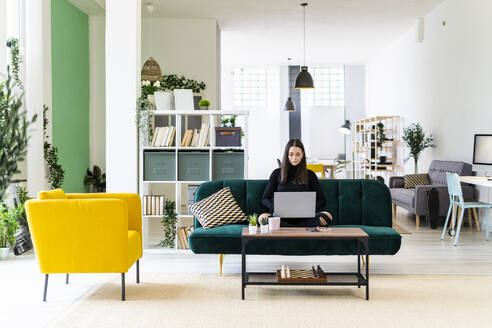 Young female influencer blogging through laptop while sitting on sofa at loft apartment - GIOF09424